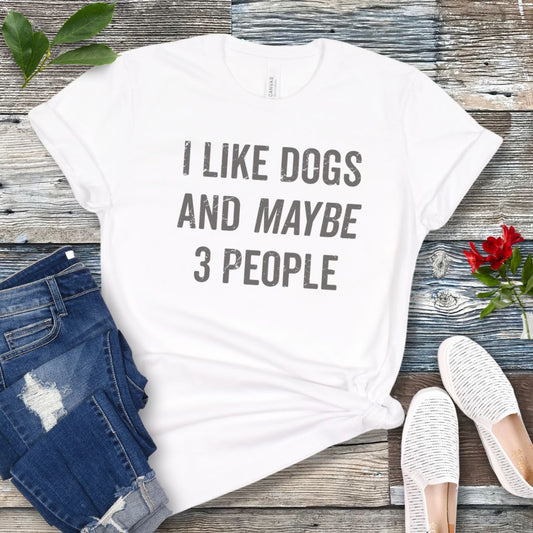 White t-shirt for dog lovers with humorous text 'I LIKE DOGS AND MAYBE 3 PEOPLE', shown on a wooden surface with jeans and shoes.