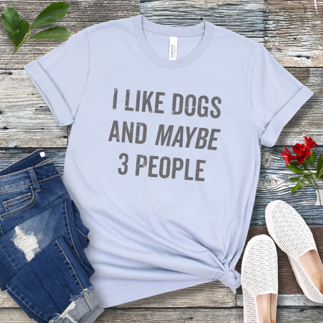 Light blue t-shirt with 'I LIKE DOGS AND MAYBE 3 PEOPLE' text on a wooden surface with jeans and shoes.