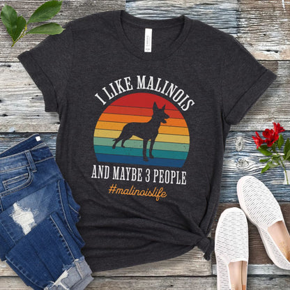 Dark grey t-shirt with Belgian Malinois dog graphic, vintage sunset and the text 'I like Malinois and maybe 3 people. #malinoislife', presented on a wooden surface with jeans and shoes.