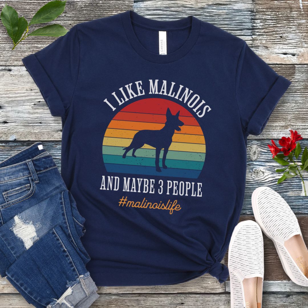 Navy blue t-shirt with Belgian Malinois dog graphic, vintage sunset and the text 'I like Malinois and maybe 3 people. #malinoislife', presented on a wooden surface with jeans and shoes.