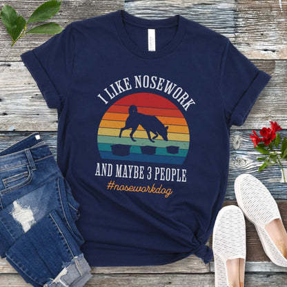 Navy blue nosework t-shirt with sniffing dog graphic and "I Like Nosework and Maybe 3 People" text on a wooden surface with jeans and shoes.