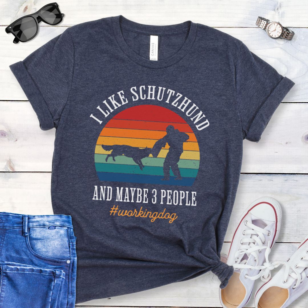 Heather navy t-shirt with vintage sunset, bitework dog sport graphic, and the text 'I like Schutzhund and maybe 3 people, #workingdog', presented on a wooden surface with jeans and shoes.