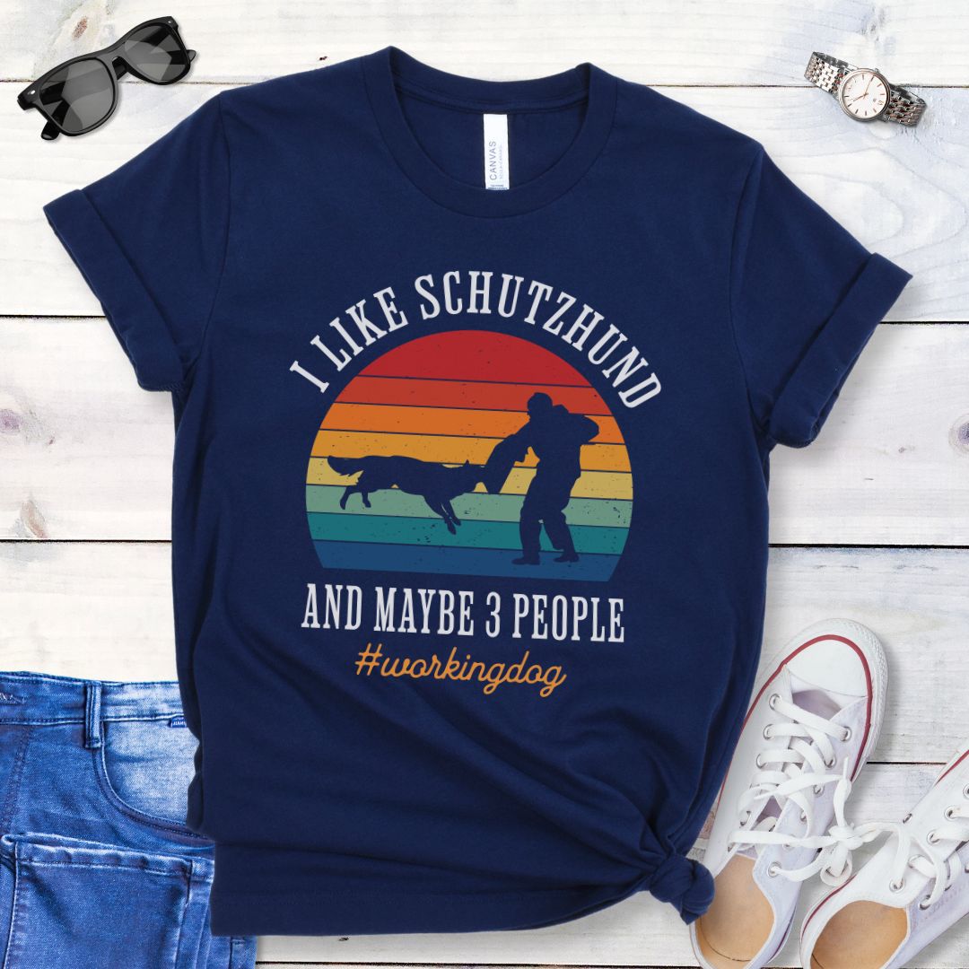 Navy blue t-shirt with vintage sunset, bitework dog sport graphic, and the text 'I like Schutzhund and maybe 3 people, #workingdog', presented on a wooden surface with jeans and shoes.