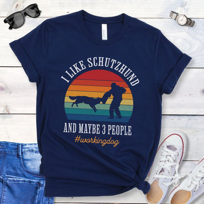 Navy blue t-shirt with vintage sunset, bitework dog sport graphic, and the text 'I like Schutzhund and maybe 3 people, #workingdog', presented on a wooden surface with jeans and shoes.