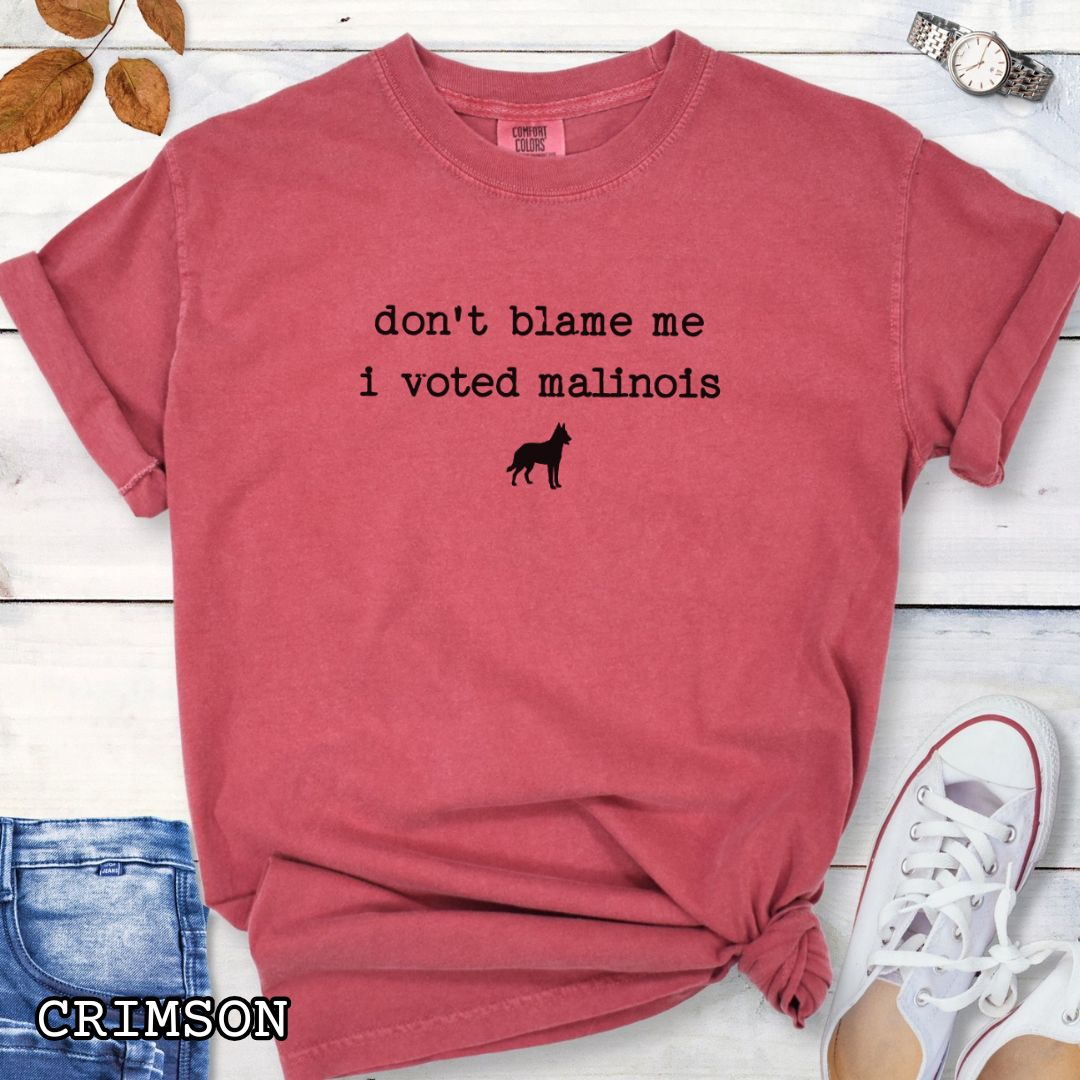 Red Comfort Colors t-shirt for Belgian Malinois lovers with humorous text, 'don't blame me, i voted malinois' and dog graphic, shown on a wooden surface with accessories.