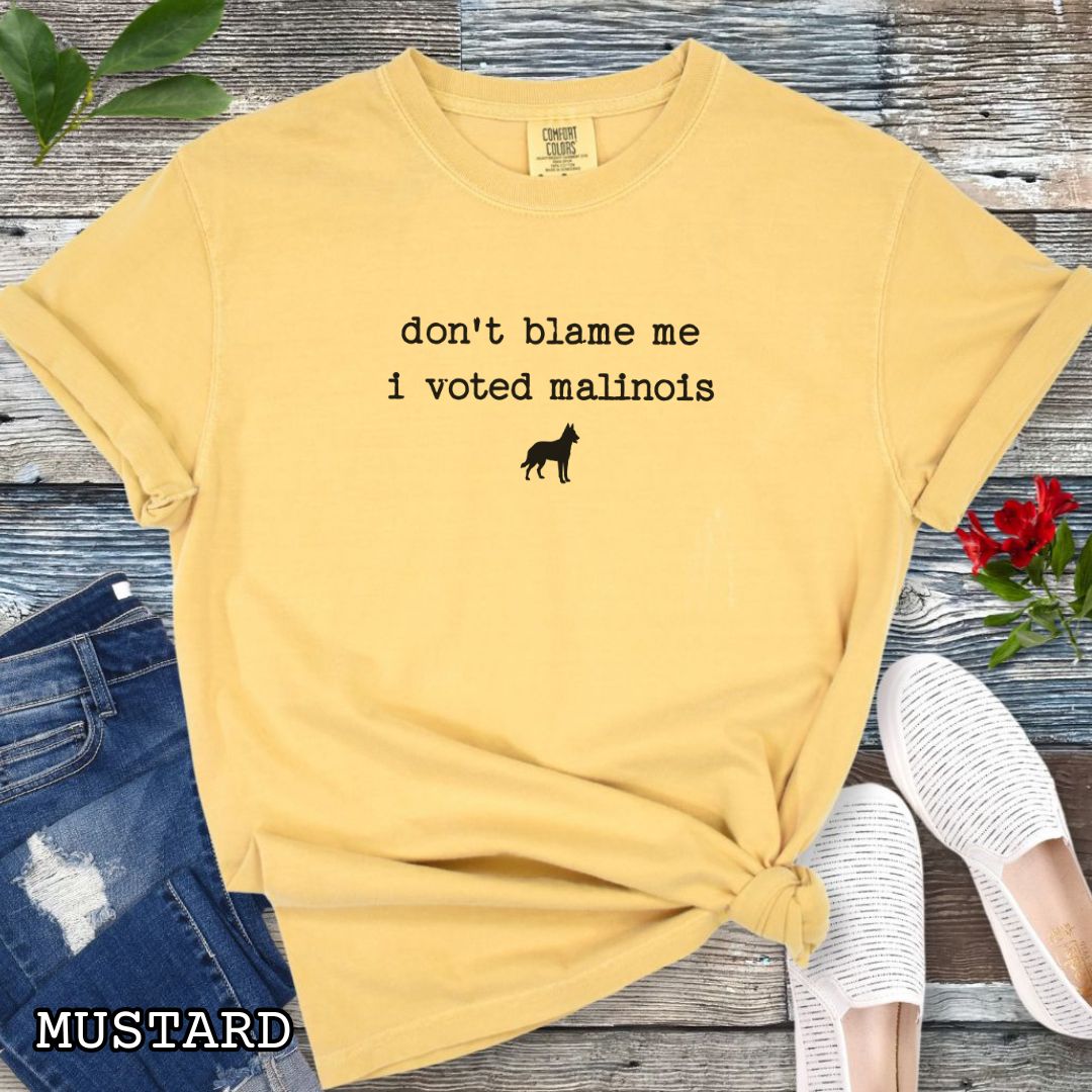 Mustard yellow Comfort Colors t-shirt for Belgian Malinois lovers with humorous text, 'don't blame me, i voted malinois' and dog graphic, shown on a wooden surface with accessories.