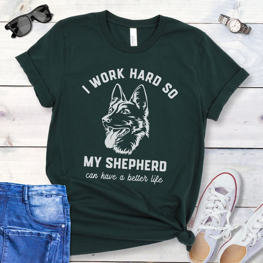 Dark Green unisex t-shirt for German Shepherd lovers with GSD graphic and humorous text 'I work hard so my shepherd can have a better life', shown on a wooden surface with jeans and shoes. 