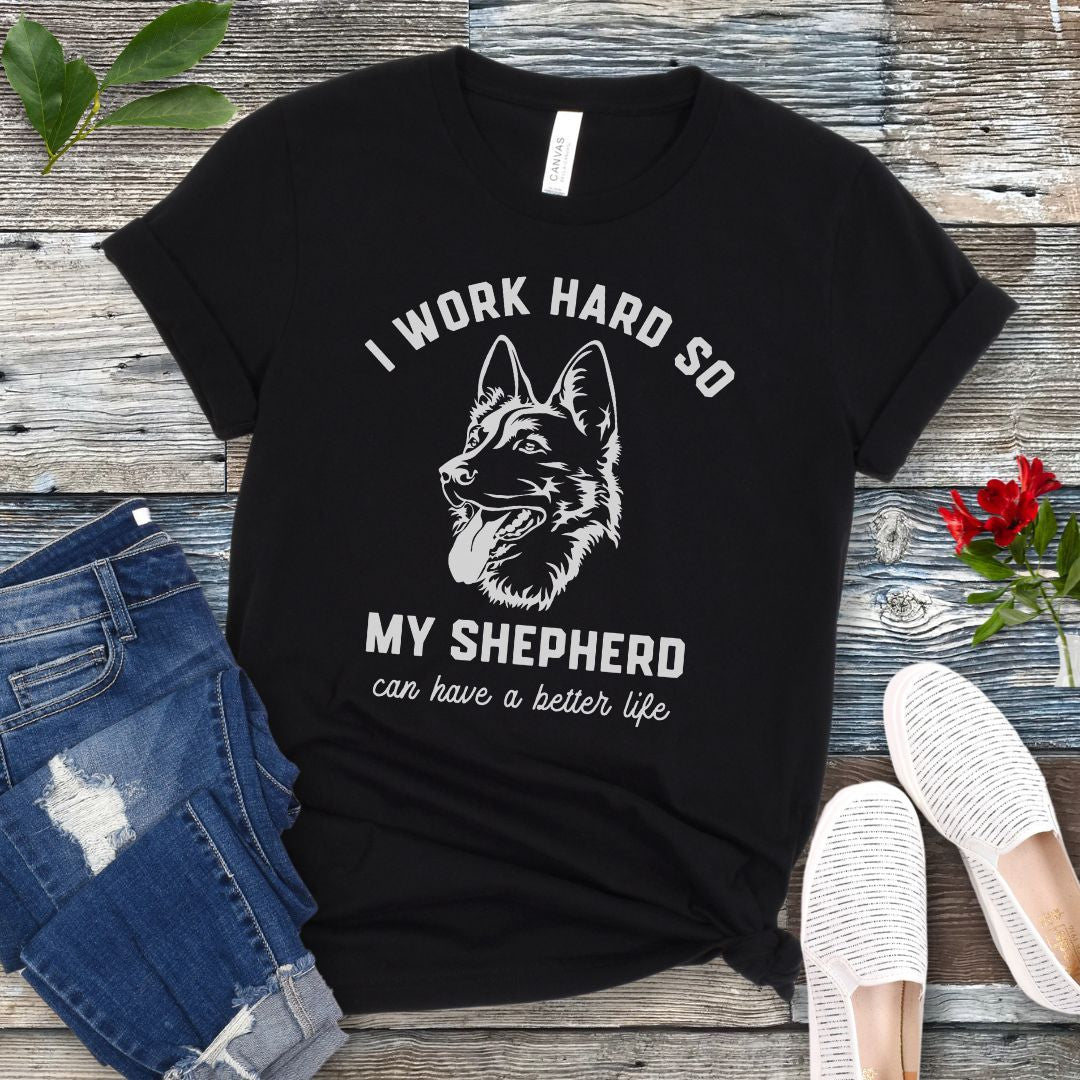 Black unisex t-shirt for German Shepherd lovers with GSD graphic and humorous text 'I work hard so my shepherd can have a better life', shown on a wooden surface with jeans and shoes. 