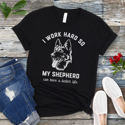 Black unisex t-shirt for German Shepherd lovers with GSD graphic and humorous text 'I work hard so my shepherd can have a better life', shown on a wooden surface with jeans and shoes. 