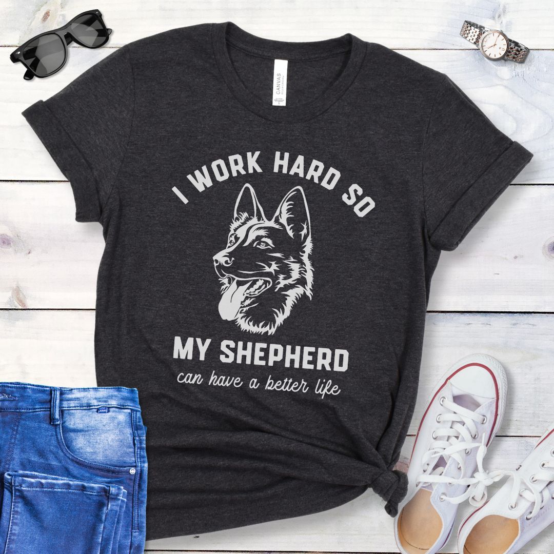 Dark Grey unisex t-shirt for German Shepherd lovers with GSD graphic and humorous text 'I work hard so my shepherd can have a better life', shown on a wooden surface with jeans and shoes. 