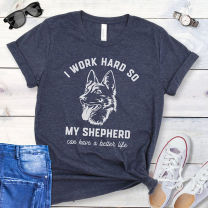 Heather navy blue unisex t-shirt for German Shepherd lovers with GSD graphic and humorous text 'I work hard so my shepherd can have a better life', shown on a wooden surface with jeans and shoes. 