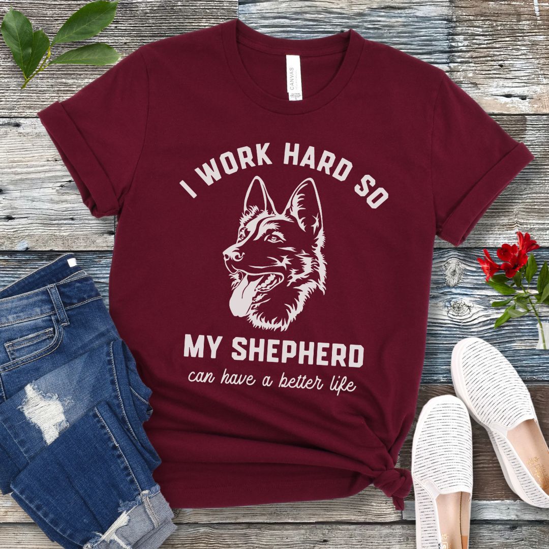 Maroon unisex t-shirt for German Shepherd lovers with GSD graphic and humorous text 'I work hard so my shepherd can have a better life', shown on a wooden surface with jeans and shoes. 