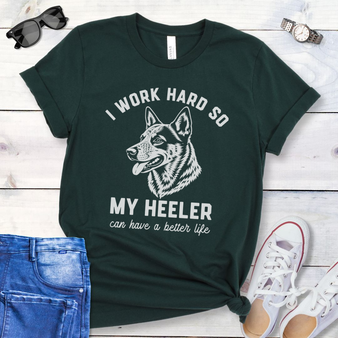 Dark green unisex t-shirt for Australian Cattle Dog lovers with linocut-style Heeler head study graphic and humorous text 'I work hard so my Heeler can have a better life', shown on a wooden surface with jeans and shoes. 