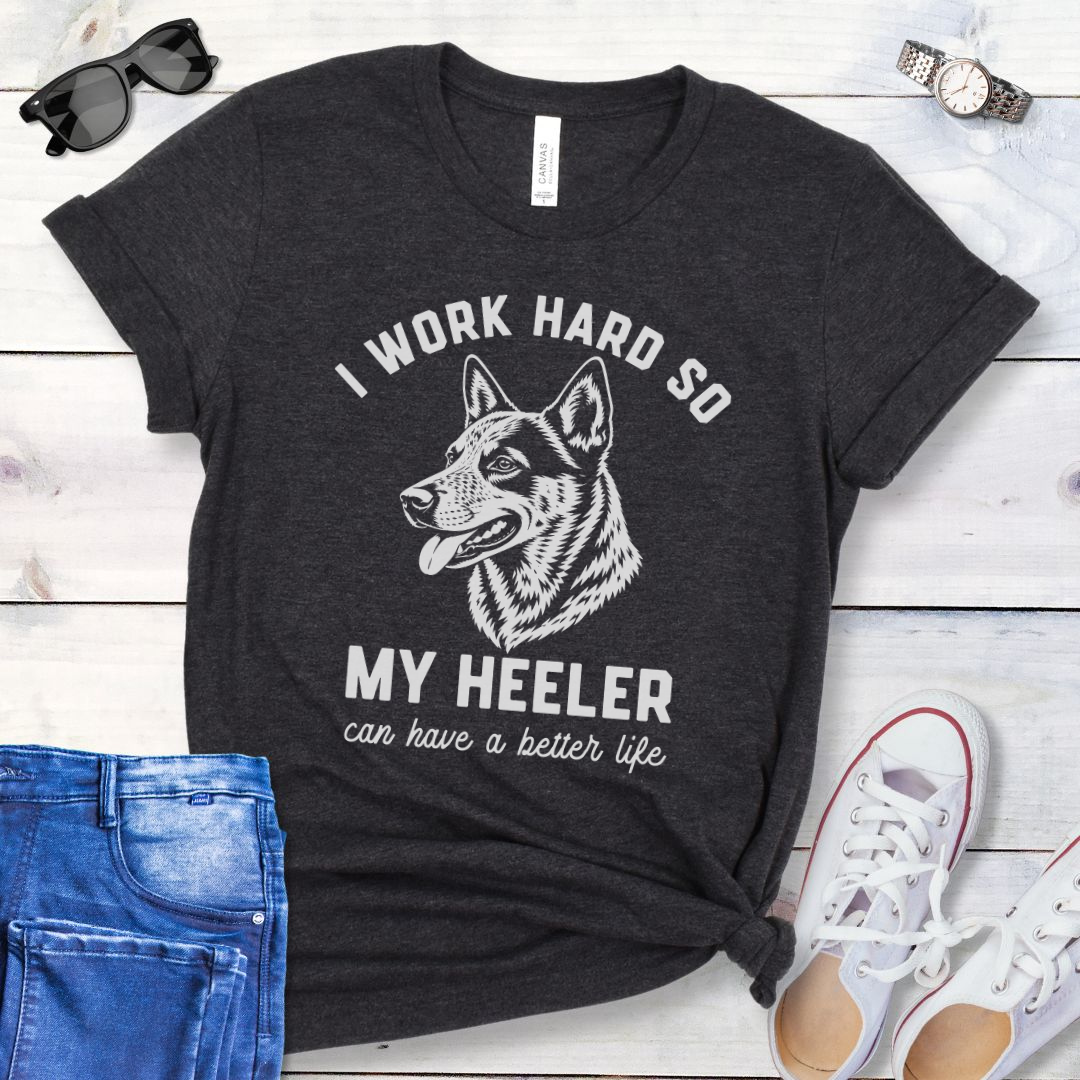 Dark grey unisex t-shirt for Australian Cattle Dog lovers with linocut-style Heeler head study graphic and humorous text 'I work hard so my Heeler can have a better life', shown on a wooden surface with jeans and shoes. 
