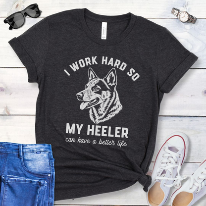 Dark grey unisex t-shirt for Australian Cattle Dog lovers with linocut-style Heeler head study graphic and humorous text 'I work hard so my Heeler can have a better life', shown on a wooden surface with jeans and shoes. 