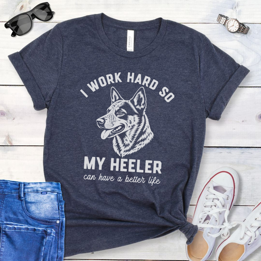 Heather navy blue unisex t-shirt for Australian Cattle Dog lovers with linocut-style Heeler head study graphic and humorous text 'I work hard so my Heeler can have a better life', shown on a wooden surface with jeans and shoes. 