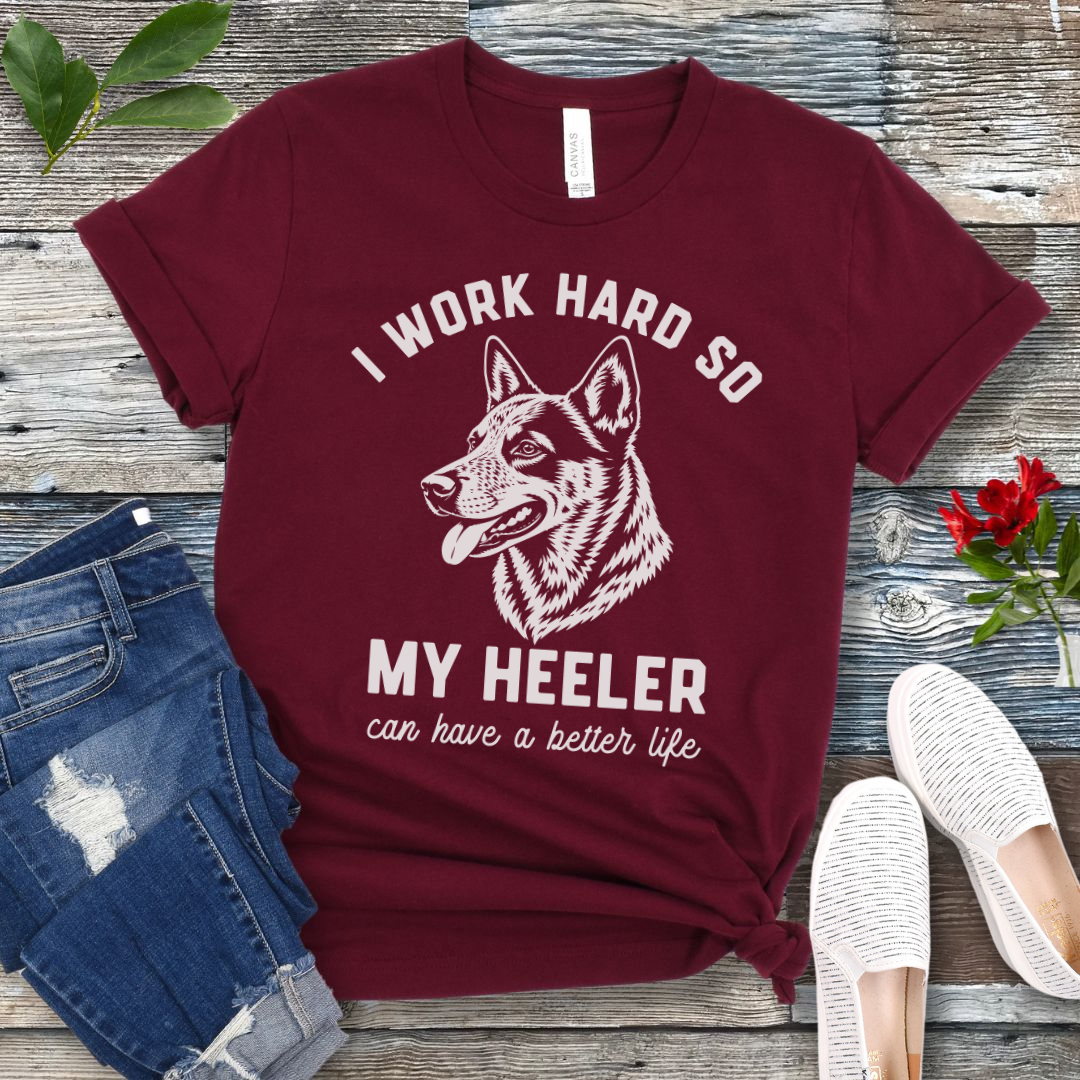 Maroon unisex t-shirt for Australian Cattle Dog lovers with linocut-style Heeler head study graphic and humorous text 'I work hard so my Heeler can have a better life', shown on a wooden surface with jeans and shoes. 