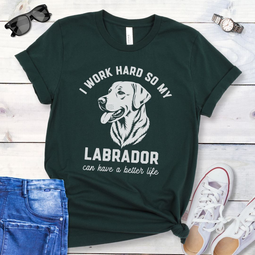 Dark green unisex t-shirt for Labrador Retriever lovers with linocut style Labrador head study graphic and humorous text 'I work hard so my Labrador can have a better life', shown on a wooden surface with jeans and shoes. 