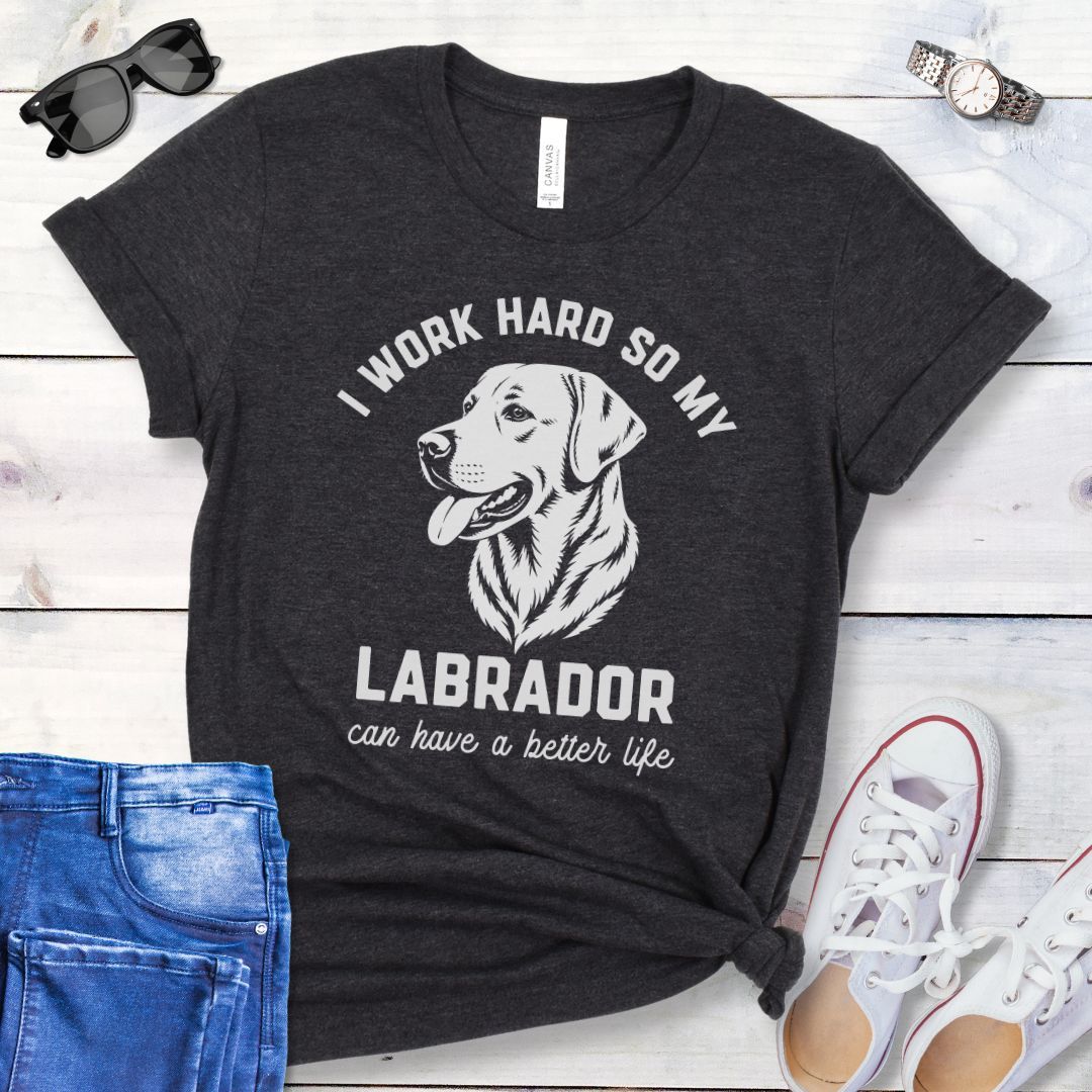 Dark grey unisex t-shirt for Labrador Retriever lovers with linocut style Labrador head study graphic and humorous text 'I work hard so my Labrador can have a better life', shown on a wooden surface with jeans and shoes. 
