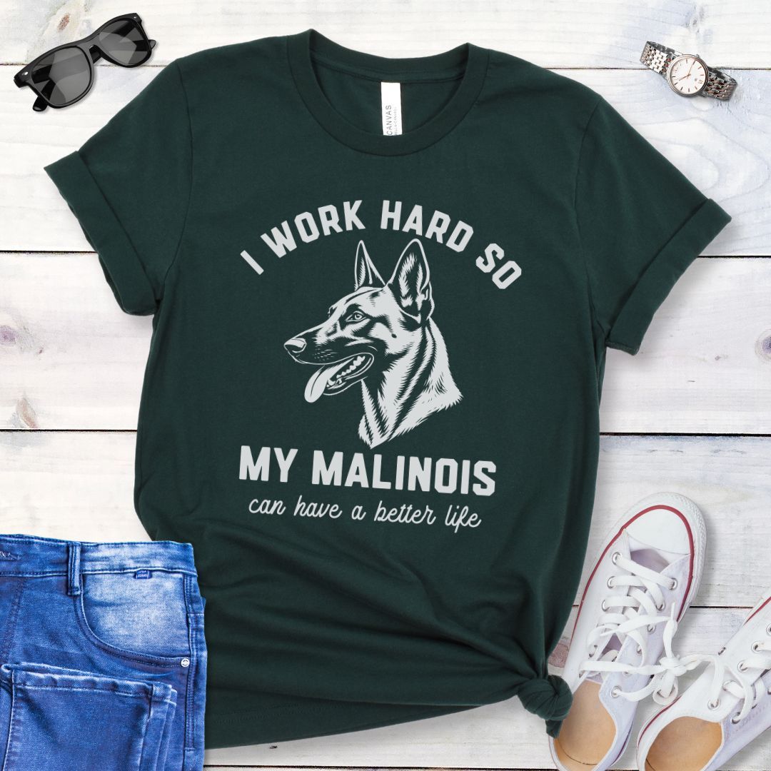 Dark green unisex t-shirt for Belgian Malinois lovers with Malinois graphic and humorous text 'I work hard so my Malinois can have a better life', shown on a wooden surface with jeans and shoes. 