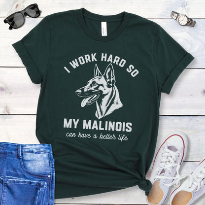 Dark green unisex t-shirt for Belgian Malinois lovers with Malinois graphic and humorous text 'I work hard so my Malinois can have a better life', shown on a wooden surface with jeans and shoes. 