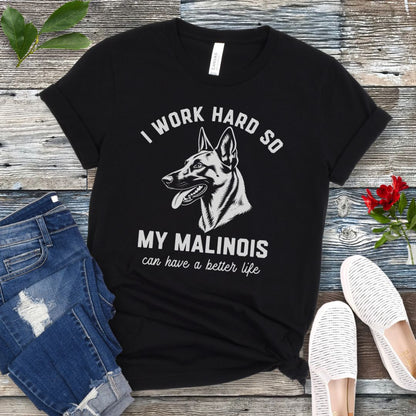 Black unisex t-shirt for Belgian Malinois lovers with Malinois graphic and humorous text 'I work hard so my Malinois can have a better life', shown on a wooden surface with jeans and shoes. 