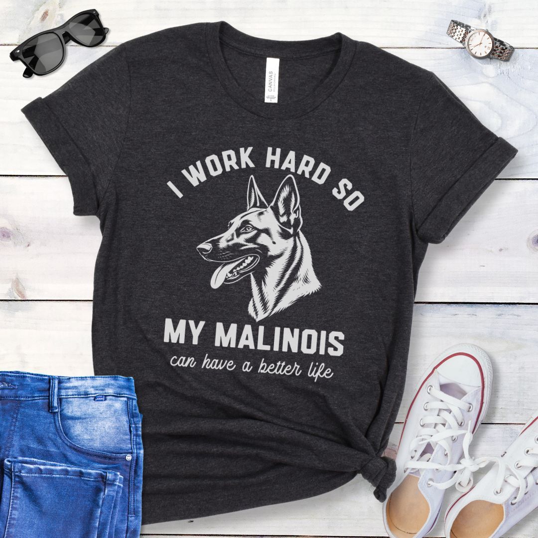 Dark grey unisex t-shirt for Belgian Malinois lovers with Malinois graphic and humorous text 'I work hard so my Malinois can have a better life', shown on a wooden surface with jeans and shoes. 