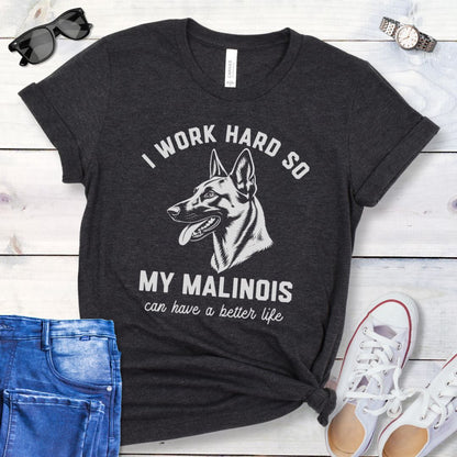 Dark grey unisex t-shirt for Belgian Malinois lovers with Malinois graphic and humorous text 'I work hard so my Malinois can have a better life', shown on a wooden surface with jeans and shoes. 