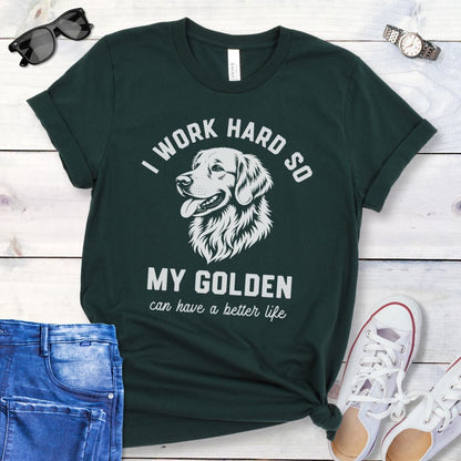 Dark green unisex t-shirt for Golden Retriever lovers with linocut-style breed head study graphic and humorous text 'I work hard so my Golden can have a better life', shown on a wooden surface with jeans and shoes. 