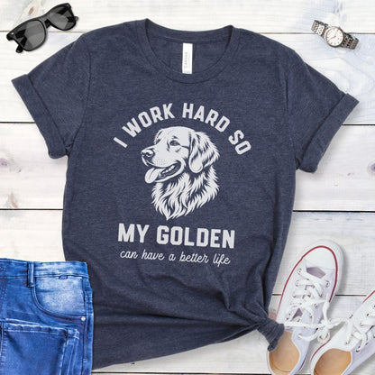 Heather navy blue unisex t-shirt for Golden Retriever lovers with linocut-style breed head study graphic and humorous text 'I work hard so my Golden can have a better life', shown on a wooden surface with jeans and shoes. 