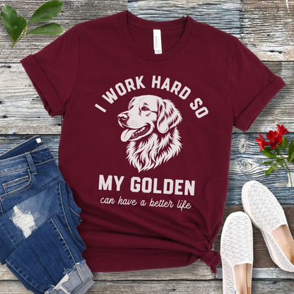 Maroon unisex t-shirt for Golden Retriever lovers with linocut-style breed head study graphic and humorous text 'I work hard so my Golden can have a better life', shown on a wooden surface with jeans and shoes. 