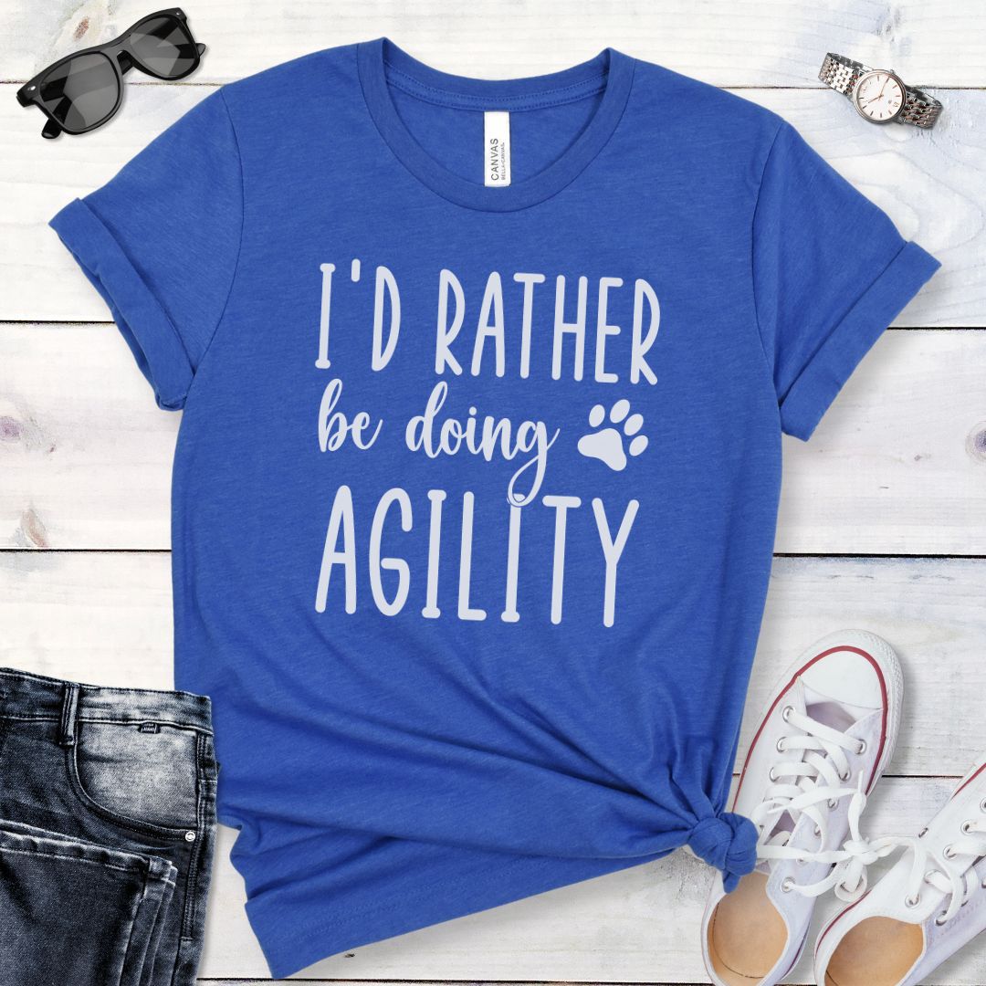 Royal blue dog sports t-shirt with 'I'd Rather be doing Agility' text on a wooden surface with jeans and shoes.