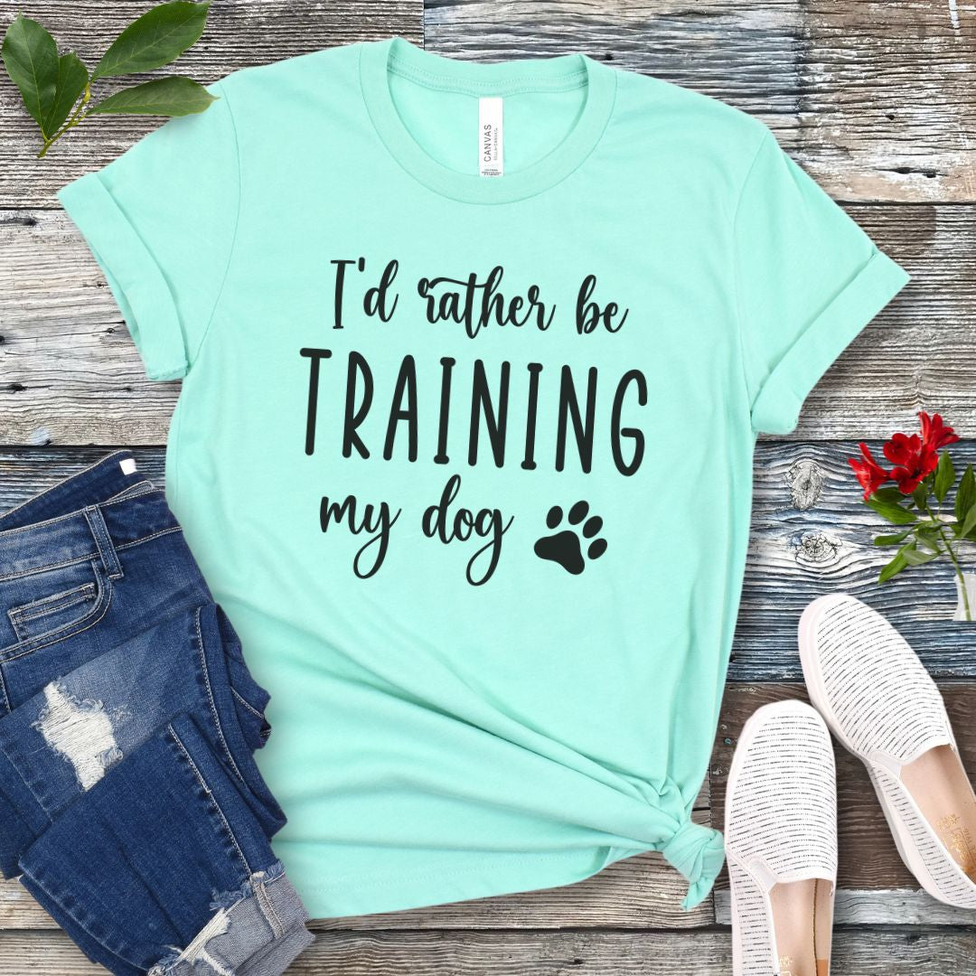 Mint green unisex t-shirt with 'I'd rather be training my dog' text in playful, feminine font with paw print, presented on a wooden surface with jeans and shoes.