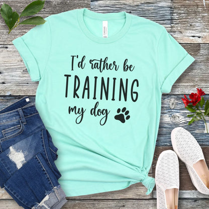 Mint green unisex t-shirt with 'I'd rather be training my dog' text in playful, feminine font with paw print, presented on a wooden surface with jeans and shoes.