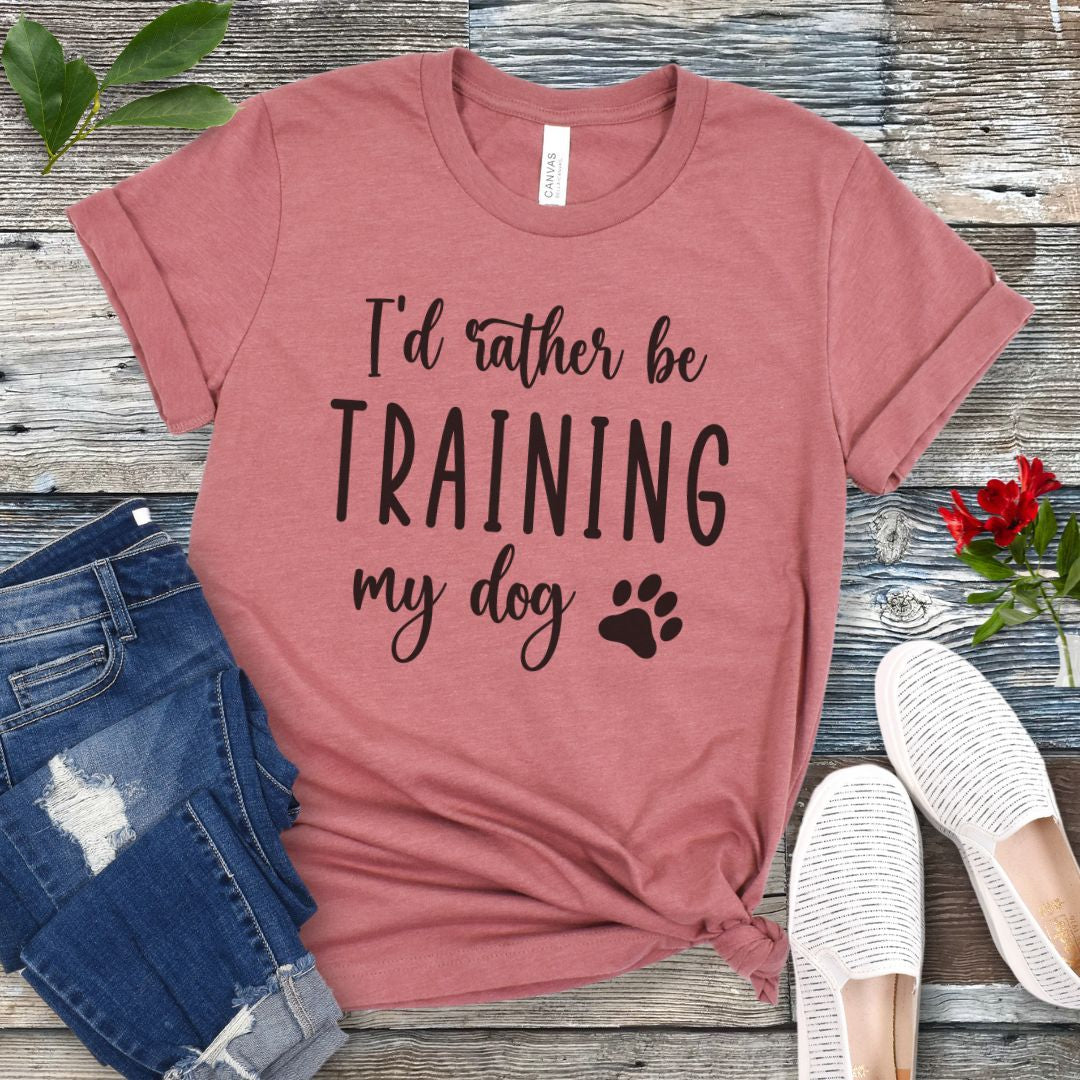 Mauve unisex t-shirt with 'I'd rather be training my dog' text in playful, feminine font with paw print, presented on a wooden surface with jeans and shoes.
