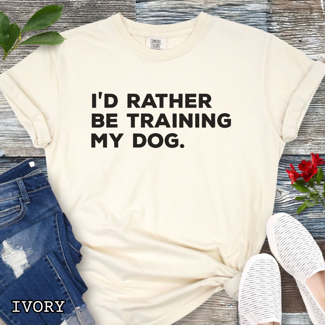 Beige (Ivory) Comfort Colors t-shirt with 'I'd rather be training my dog' text on a light wooden surface with shoes and jeans.