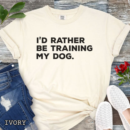 Beige (Ivory) Comfort Colors t-shirt with 'I'd rather be training my dog' text on a light wooden surface with shoes and jeans.