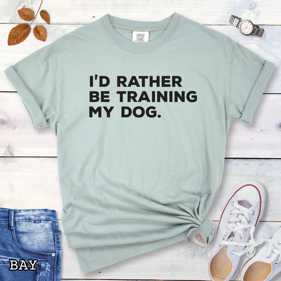 Light green (Bay) Comfort Colors t-shirt with 'I'd rather be training my dog' text on a light wooden surface with shoes and jeans.