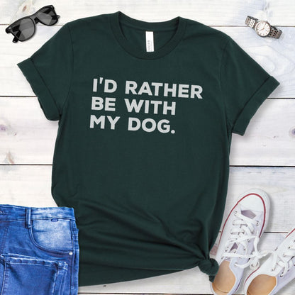 Dark green t-shirt for dog lovers with 'I'd Rather Be With My Dog' text in bold lettering, shown on a wooden surface with jeans and shoes.