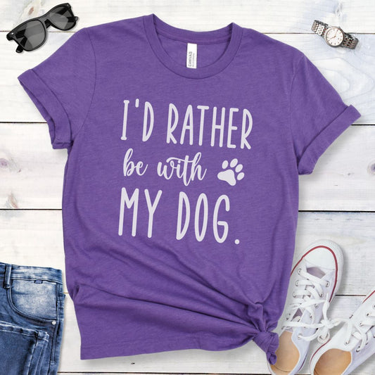 Purple t-shirt for dog lovers and dog moms with the humorous text 'I'd rather be with my dog' in playful, handwritten font, shown on a wooden surface with jeans and shoes.