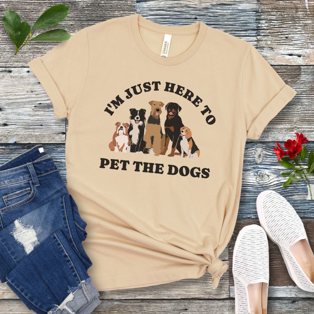 A cream colored t-shirt for dog lovers with the text 'I'M JUST HERE TO PET THE DOGS' accompanied by an image of five dogs, displayed on a wooden surface next to a pair of blue jeans and white shoes.