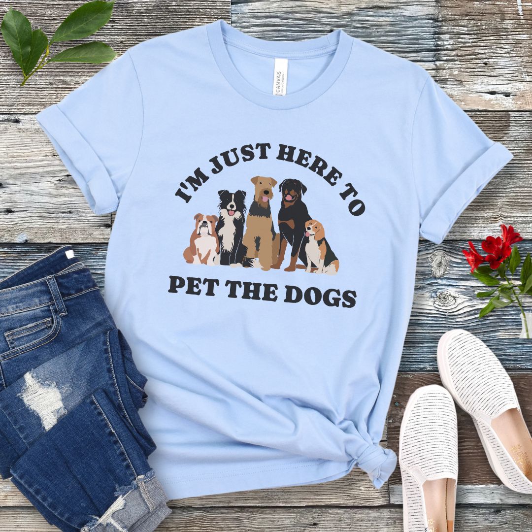 A light blue t-shirt for dog lovers with the text 'I'M JUST HERE TO PET THE DOGS' accompanied by an image of five dogs, displayed on a wooden surface next to a pair of blue jeans and white shoes.