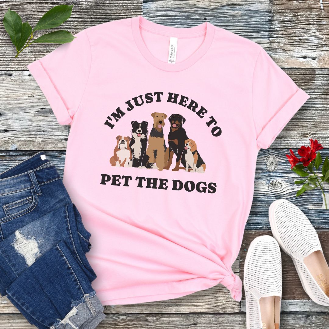 A pink t-shirt for dog lovers with the text 'I'M JUST HERE TO PET THE DOGS' accompanied by an image of five dogs, displayed on a wooden surface next to a pair of blue jeans and white shoes.
