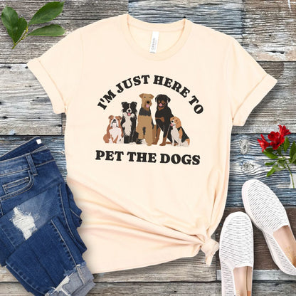 A beige colored t-shirt for dog lovers with the text 'I'M JUST HERE TO PET THE DOGS' accompanied by an image of five dogs, displayed on a wooden surface next to a pair of blue jeans and white shoes.