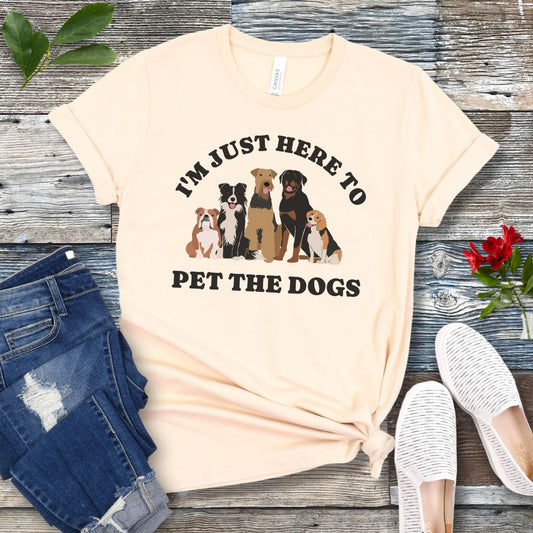 A beige colored t-shirt for dog lovers with the text 'I'M JUST HERE TO PET THE DOGS' accompanied by an image of five dogs, displayed on a wooden surface next to a pair of blue jeans and white shoes.