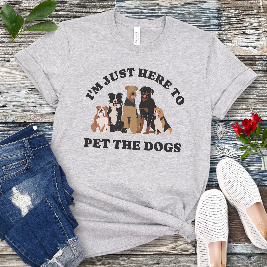 A light grey t-shirt for dog lovers with the text 'I'M JUST HERE TO PET THE DOGS' accompanied by an image of five dogs, displayed on a wooden surface next to a pair of blue jeans and white shoes.