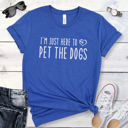 A royal blue t-shirt for dog lovers with the text 'I'M JUST HERE TO PET THE DOGS' printed in playful text with paw-in-heart graphic, displayed flat with a pair of rolled-up blue jeans and white shoes.