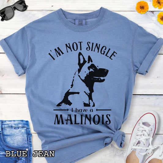 Blue Comfort Colors t-shirt with a woodcut-style Belgian Malinois graphic and the humorous text 'I'm Not Single, I Have a Malinois', shown on a wooden surface with accessories.