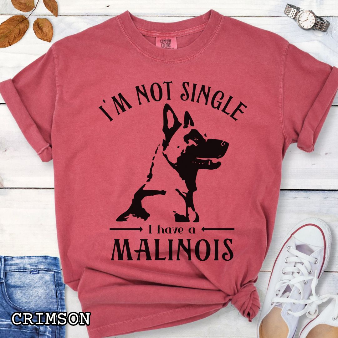 Red Comfort Colors t-shirt with a woodcut-style Belgian Malinois graphic and the humorous text 'I'm Not Single, I Have a Malinois', shown on a wooden surface with accessories.