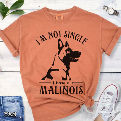 Orange Comfort Colors t-shirt with a woodcut-style Belgian Malinois graphic and the humorous text 'I'm Not Single, I Have a Malinois', shown on a wooden surface with accessories.